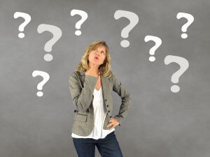 A woman with long blonde hair stands pensively, her right hand touching her chin. She is wearing a grey blazer over a white blouse and dark jeans. Surrounded by large white question marks on a grey background, she appears to be contemplating important decisions.