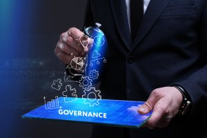 A person in a suit holds a transparent tablet with the word "GOVERNANCE" on it. Holographic icons representing ideas, money, gears, and analytics float above the tablet, symbolizing various aspects of governance and business strategy in the post-COVID-19 world.