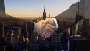Double exposure image of a city skyline at sunset with two businesspeople shaking hands in the foreground. The cityscape includes tall skyscrapers, and the handshake symbolizes a business agreement or partnership. The tones are warm and professional.