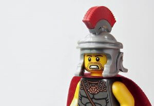 Close-up of a Lego minifigure dressed as a Roman soldier, embodying crisis leadership. The figure wears a grey helmet with a red plume, detailed chest armor, and a red cape. With a stern expression and bearded face against a plain white background, it exudes command and resilience.