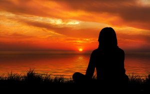 A silhouette of a person sitting cross-legged on the grass, perhaps taking serene naps, gazes at a vibrant sunset over a calm body of water. The sky is filled with warm hues of orange, red, and yellow, with scattered clouds adding to the picturesque scene.