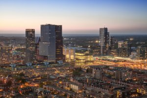 Aerial view of a cityscape at sunset featuring modern skyscrapers, residential buildings, and a well-lit train station. The sky transitions from light to dark blue, with streetlights and buildings illuminating the scene below, highlighting the city's commitment to public service infrastructure.