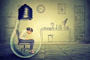 A woman sits in a chair while working on a laptop inside a giant light bulb. The background features a hand-drawn style office with a clock, bookshelves, desks, and computer. The overall image depicts the creative workspace concept essential for effective crisis leadership.