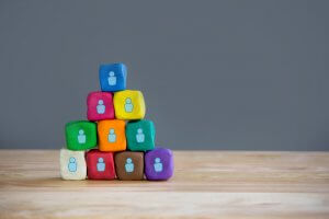 A pyramid of colorful clay blocks sits on a wooden surface against a gray background. Each block displays a white icon of a person, symbolizing unity during COVID-19. The pyramid consists of ten blocks in various colors such as blue, yellow, green, red, orange, purple, brown, and teal.