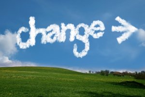 A field of green grass under a clear blue sky with a few trees in the distance. In the sky, clouds are shaped to spell the word "change" followed by an arrow pointing to the right, hinting at shifting weather patterns.