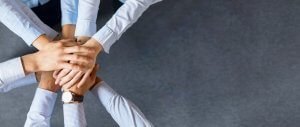 A group of people in business attire stand in a circle, placing their hands on top of one another in a display of pandemic leadership and unity. Their sleeves are mostly light colors, and one person is wearing a watch. The background is a plain, dark grey surface.