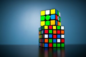 Two stacked Rubik's Cubes on a dark surface against a gradient blue background. The top cube has some solved sides, while the bottom cube is partially solved, with mixed colored squares visible. The cubes are brightly lit, emphasizing their vivid colors and the challenge to solve problems.