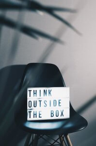A black chair is placed against a grey wall, with a lightbox sign on the seat displaying the phrase "THINK OUTSIDE THE BOX." Blurred plant leaves are partially visible in the foreground, subtly suggesting a fresh perspective to solve problems.