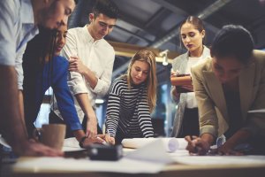 A group of six people is gathered around a table, actively engaged in discussing and working on documents. Some are standing, while others are leaning over the table, collaborating on ideas to solve problems in a modern office setting with a warm ambiance.