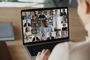A person is participating in a video conference on a laptop, showing 12 small screens of diverse individuals in a grid format. The participants are engaging, smiling, and appear to be discussing COVID-19 staffing solutions. There are bookshelves and home environments in the background.