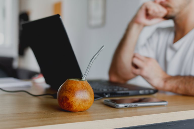 A person sits at a desk with their hand resting on their chin, focused on a laptop. In the foreground, a traditional gourd with a metal straw for drinking yerba mate is next to a smartphone. The scene suggests remote work or study during coronavirus confinement.