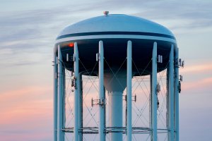 A tall, white water tower with a large blue and white tank on top, supported by multiple columns. Attached to the columns are various antennas and equipment essential for the water workforce. The background features a colorful sky at dusk with hues of pink, purple, and blue.