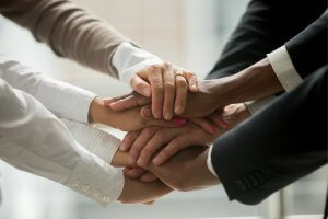A close-up of multiple hands stacked on top of each other, symbolizing unity and teamwork. The hands belong to individuals of diverse backgrounds, including both men and women, wearing professional attire such as suits and dress shirts—capturing the essence of humanity working together.