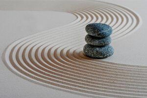 Three stacked smooth stones rest on a sand surface with curved, raked lines, creating a serene and balanced scene symbolizing Zen and tranquility. The lines form flowing patterns around the stones, enhancing the sense of calm, harmony, and overall wellness.