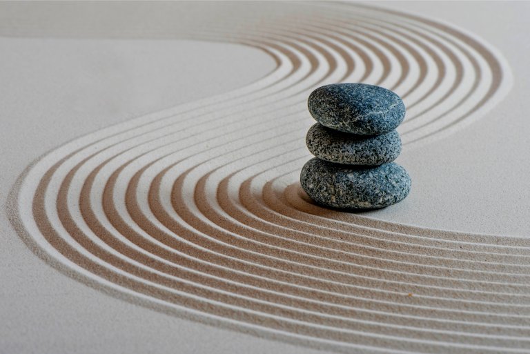 Three stacked smooth stones rest on a sand surface with curved, raked lines, creating a serene and balanced scene symbolizing Zen and tranquility. The lines form flowing patterns around the stones, enhancing the sense of calm, harmony, and overall wellness.