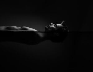 Black and white image of a person floating horizontally with their face just above the waterline, creating a serene and reflective atmosphere. The darkness frames the subject's silhouette, emphasizing both the calmness and isolation of the scene.