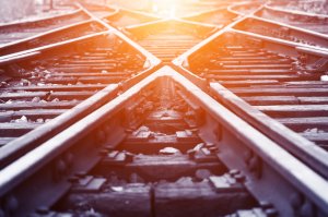 Close-up view of intersecting railway tracks converging towards a distant point, bathed in warm sunlight. The perspective creates a visual effect of the tracks forming an 'X', with wooden sleepers and pebbles visible between the rails, evoking a sense of isolation.