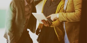 Three people in business casual attire are gathered closely, viewing and discussing something on a tablet. Their faces are not visible, with focus on the tablet and their hand gestures. They appear to be collaborating in an indoor setting, likely working on an HR project.