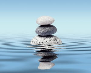 A stack of three smooth stones, in ascending size, balances atop each other in serene isolation on a calm water surface with gentle ripples. The stones are reflected perfectly in the water, and the background is a clear blue sky.