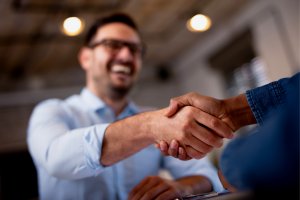 A close-up shot of two people shaking hands during an interview. One person's face is visible and smiling, while the other person's face is out of frame. The background is slightly blurred, indicating an indoor setting, possibly an office.