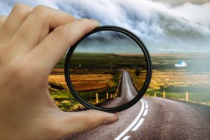 A hand holds a circular camera filter over a landscape, aligning it with a road that stretches into the distance. The filter brings clarity to the scene, creating a clearer, more vivid view of the road, contrasting with the slightly blurred and less vibrant background.
