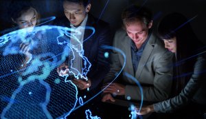 A group of four people are gathered around a table in a dark room, illuminated by a holographic projection of a world map. They appear to be interacting with the hologram, looking engaged and focused as they discuss its influences on global trends. Blue and white lights from the hologram cast a glow on their faces.