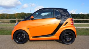 An orange Smart car with black detailing, parked on a paved surface, showcases influences of sleek modern design. The compact two-door vehicle is viewed from the side, with a wooden fence and grassy area with trees in the background under a partly cloudy blue sky.