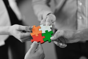 Four hands each hold a piece of a jigsaw puzzle, joining them together in a display of teamwork. The puzzle pieces are in vibrant colors: orange, yellow, red, and green. The background is blurred and in black and white, emphasizing the colorful puzzle pieces and cooperative hands.