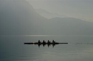 Four people are rowing a boat on a calm body of water with mist-covered mountains in the background. Their synchronized teamwork, combined with the soft lighting and fog, creates a serene and peaceful atmosphere.
