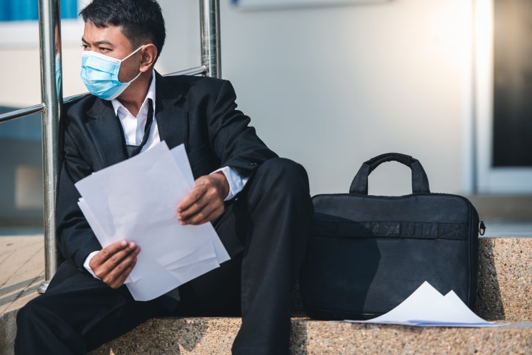 A man in a suit and a blue surgical mask sits on outdoor steps, holding papers in one hand. A black briefcase and additional papers lie next to him. He appears to be looking to his left with a thoughtful, possibly stressed, expression.