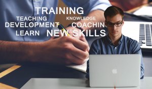 A man wearing glasses is focused on using a laptop. Overlaid around his image are words like "TRAINING," "HABITS," "DEVELOPMENT," and "COACHING." In the background, a hand is writing in a notebook with a laptop beside it.