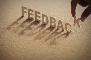 A person's hand places the letter "K" at the end of the word "FEEDBACK," enhancing its meaning on the textured brown surface. The upright letters cast elongated shadows, symbolizing impactful feedback.