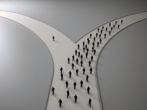 An illustration of a forked path with numerous silhouettes of people walking along the right path in unison, while one lone silhouette walks alone on the left path. The background is gradient grey, emphasizing the change between conformity and individuality.