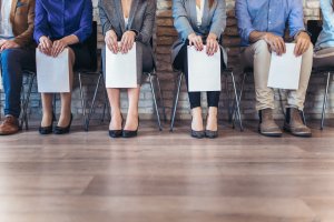 Five people are sitting in a row, holding paper documents, presumably waiting for job interviews. Only their torsos and legs are visible. They are dressed in business and business-casual attire, set against a brick wall background.