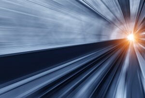 Blurred image of a tunnel, featuring dynamic blue and black lines converging towards a bright light source at the end. The scene evokes a sense of rapid movement, speed, and futuristic travel, symbolizing a transformative journey through change.