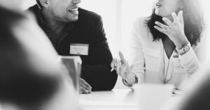 Black and white image of a man and a woman engaged in conversation at a table. The woman gestures with her hand while speaking, likely providing feedback. Both are wearing name tags, and the scene appears to be in a professional setting like a meeting or conference.