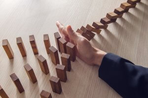A series of wooden dominoes is toppling over on a light wood surface, forming a line. A manager's hand, dressed in a dark suit sleeve, is positioned upright, halting the fall of the dominoes and preventing further collapse.