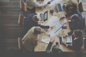 Aerial view of six people gathered around a table in a meeting. The table is filled with laptops, documents, coffee cups, and other office supplies. The manager is pointing at a blueprint while the others listen and discuss.