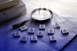 A magnifying glass is placed over an envelope on a desk, surrounded by several small wooden blocks. Each block has a simple icon of a person, symbolizing the interview selection process. Books are stacked nearby.