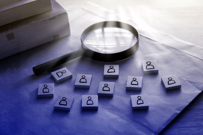A magnifying glass is placed over an envelope on a desk, surrounded by several small wooden blocks. Each block has a simple icon of a person, symbolizing the interview selection process. Books are stacked nearby.