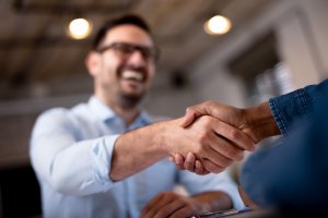 Close-up of two people shaking hands, signifying a successful interview or meeting. One person is in focus, smiling and wearing glasses. The background is slightly blurred, with visible lights and windows.