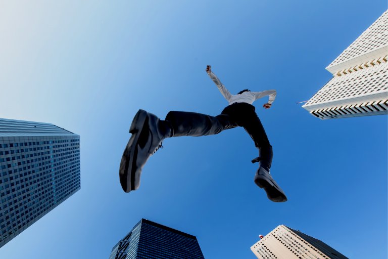 A person in business attire is captured from below, appearing to leap between tall skyscrapers. The clear blue sky emphasizes the towering buildings and the dramatic pose of the jumper, symbolizing a bold move to bridge an employment gap. The perspective provides a sense of height and motion.