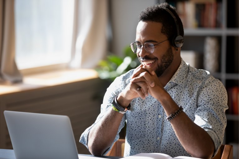 A man wearing glasses and headphones is sitting at a desk, smiling and looking at a laptop screen. Exhibiting leadership, he has his hands clasped in front of him. Bookshelves and a window with sunlight streaming in are visible in the background.