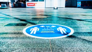 A floor sticker in a public area shows two human silhouettes with arrows between them, indicating they should stand 1.5 meters apart. The text reads "1.5M social distancing QUARANTINE." A donut shop is visible in the background.