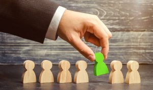 A hand in a suit selects a green wooden figurine from a row of natural wooden ones, symbolizing choice or selection during onboarding. The background is a textured wooden surface.