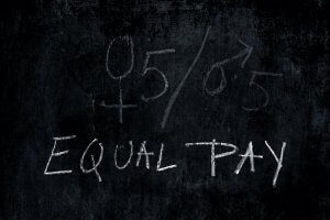 A black chalkboard has symbols for male and female side-by-side in chalk, separated by a slash, followed by "0.5/0.5." Below the symbols, the words "Equal Pay" are written in white chalk, highlighting the importance of pay equity.