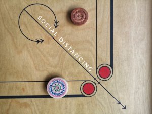 A carrom board with wooden carrom men kept apart by a diagonal line labeled "SOCIAL DISTANCING." The wooden board features black lines and red decorative circles in the corners, while one carrom man showcases colorful geometric patterns.