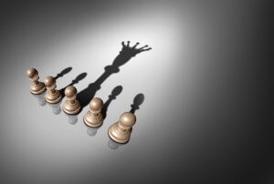 Five beige chess pawns stand in a line on a gray surface, casting shadows towards the right. Intriguingly, the shadow of the central pawn is elongated and forms the silhouette of a king piece with a crown, hinting at potential growth and indicating a defensive strategy.