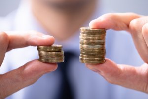 A jobseeker comparing two stacks of coins, holding a smaller stack in the left hand and a larger stack in the right hand. The background is blurred, with the person wearing a shirt and tie.