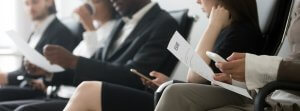 Four people sit in a row in what appears to be a waiting area or interview room. Dressed in business attire and holding resumes, they seem focused and engaged. Two of them are also using smartphones, possibly researching Government Jobs or careers in government.